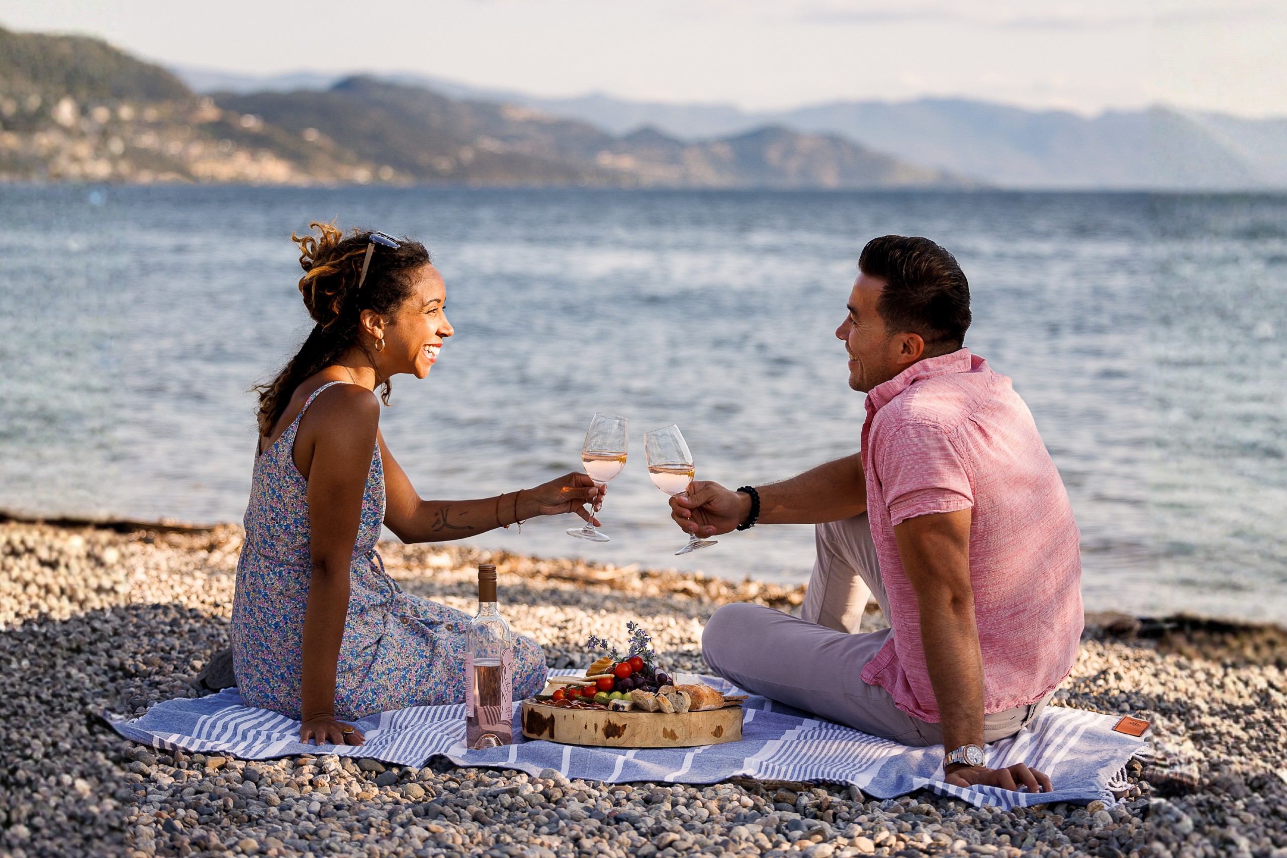 A couple smiles as their cheers their wine glasses at their beach picnic with Okanagan lake in the background.  