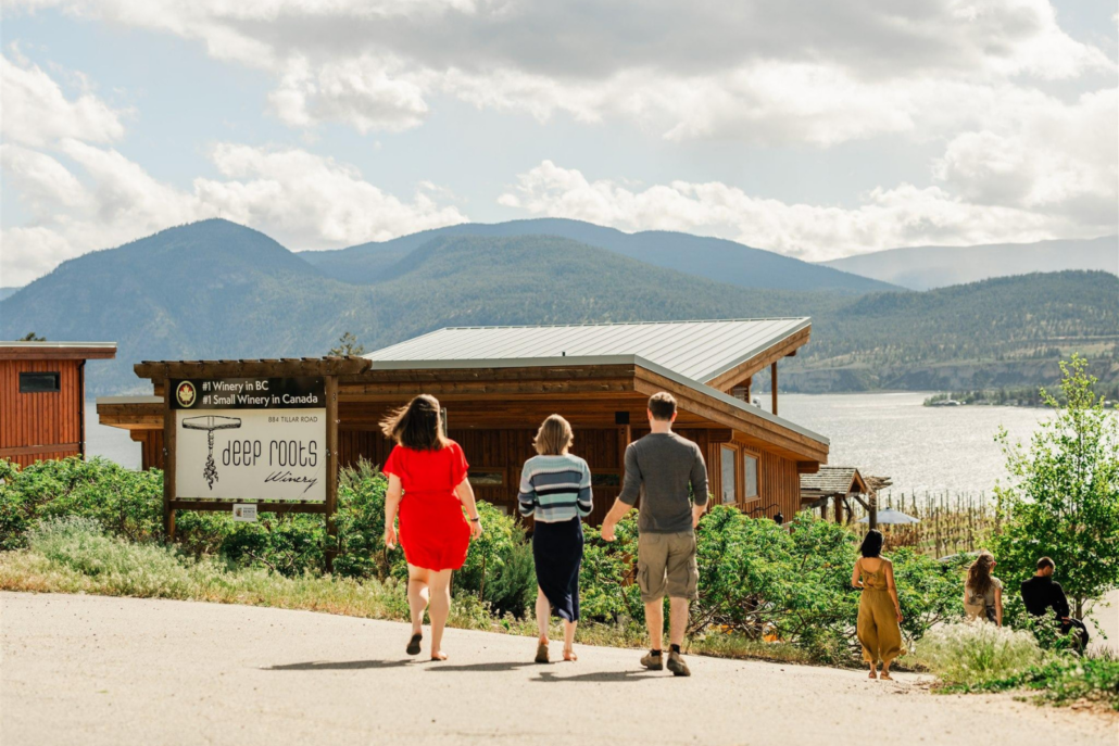 A group walking towards Deep Roots Winery with a scenic view of Okanagan Lake in the background.