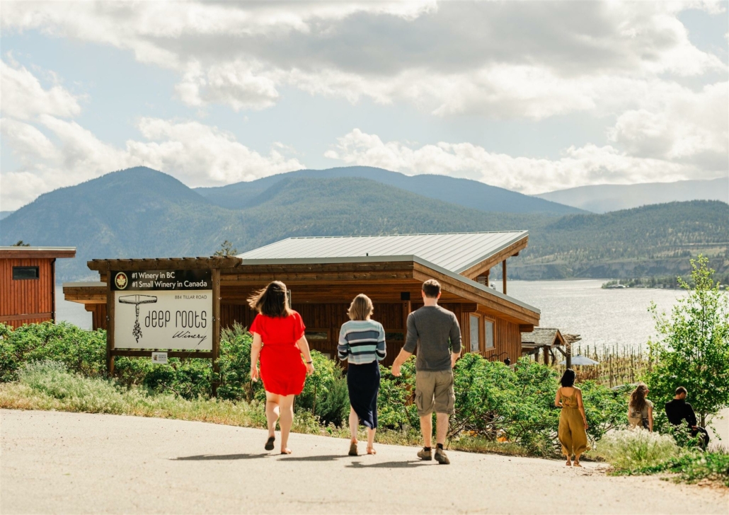 A group walking towards Deep Roots Winery with a scenic view of Okanagan Lake in the background.