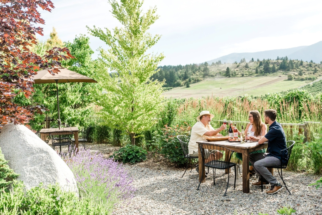 A group cheers their wine glasses on the patio at Baillie-Grohman Winery in the Kootenay's surrounded by lush garden.