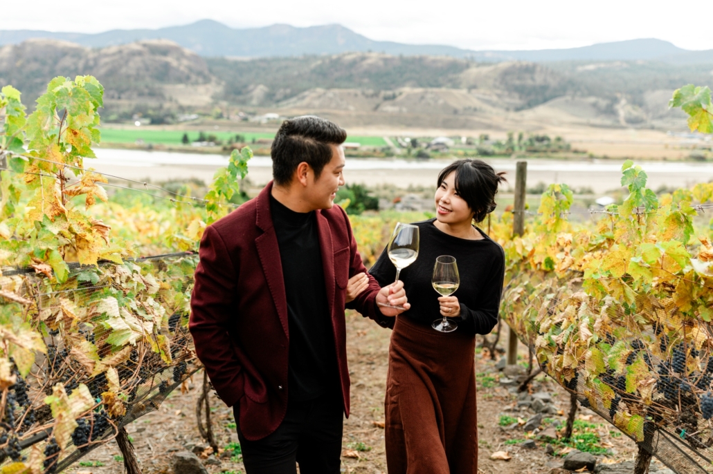 A couple walks through the colourful fall vineyard with glasses of white wine at Sagewood Winery located in Kamloops, BC.